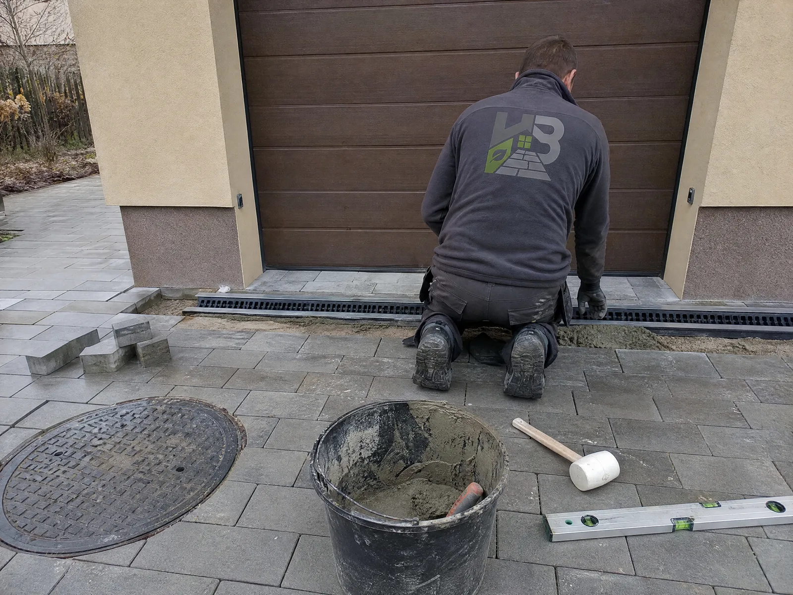 Pracownik HandyBruk osadza odwodnienie liniowe w podjeździe przed garażem, w kostce widoczna studzienka